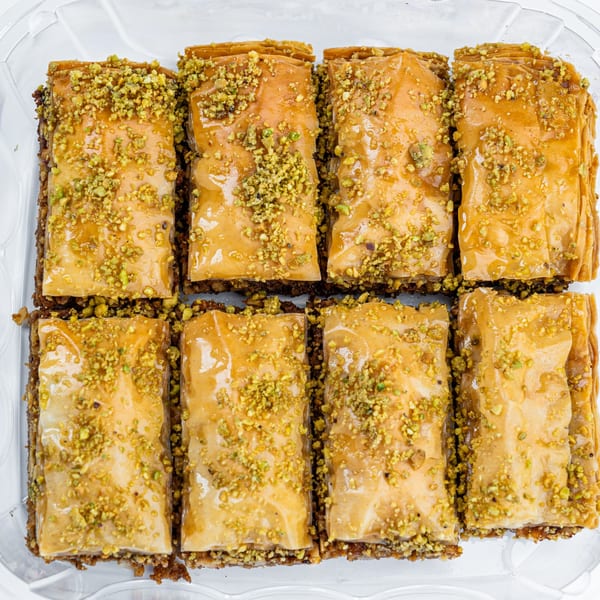 Eight pieces of golden-brown baklava, drizzled with honey and sprinkled with chopped pistachios, in a clear container.