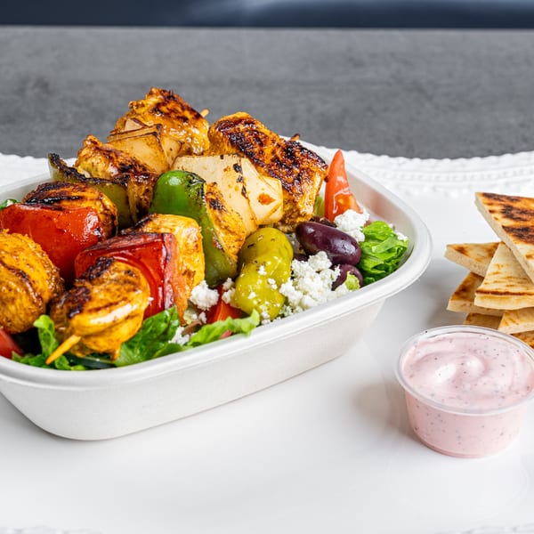 Grilled chicken and vegetable skewers over a Greek salad with pita bread and pink sauce.