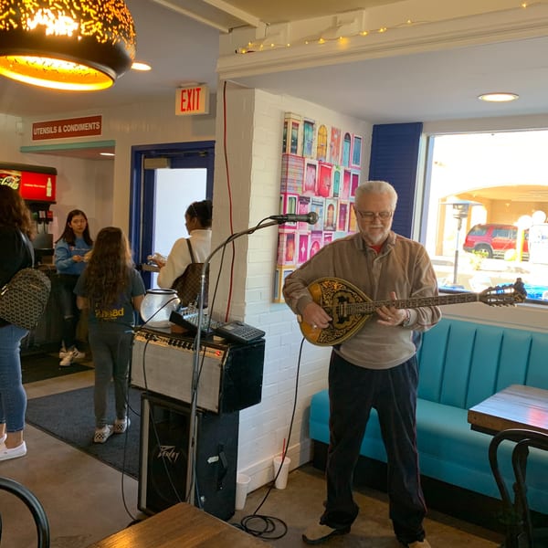 A musician plays a bouzouki in Niro's Gyros, with customers and a soda fountain in the background.